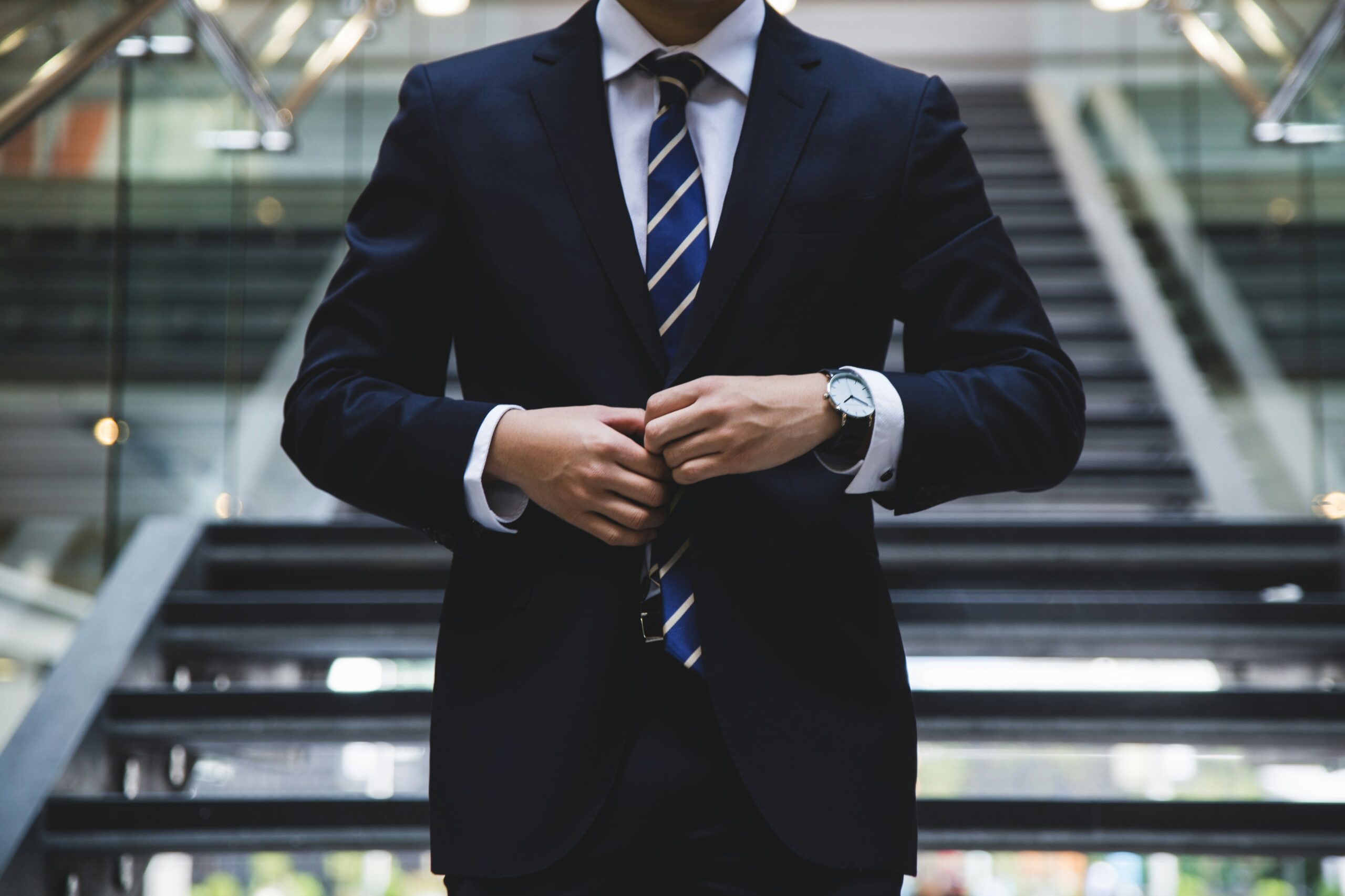 Businessman in a dark suit and striped tie adjusting his jacket, standing in front of modern office stairs, representing professionalism and corporate success.