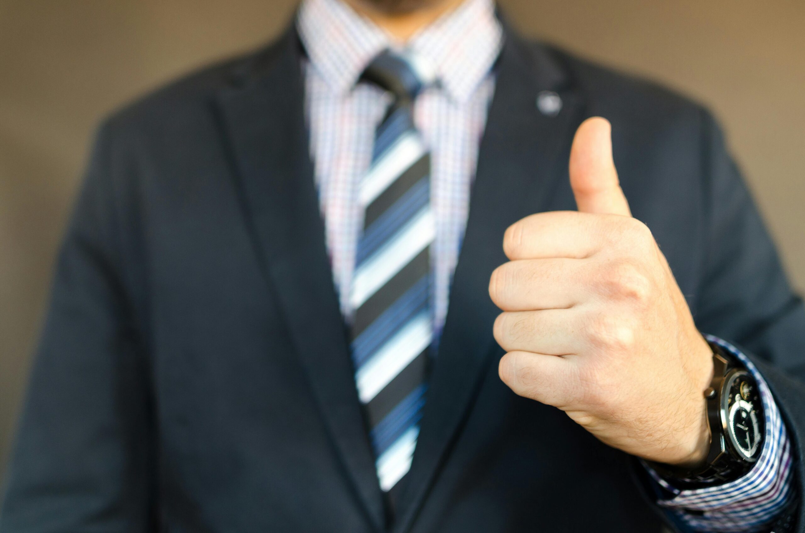 Close-up of a businessman in a suit and tie giving a thumbs-up gesture, symbolizing approval, success, and confidence in a professional setting