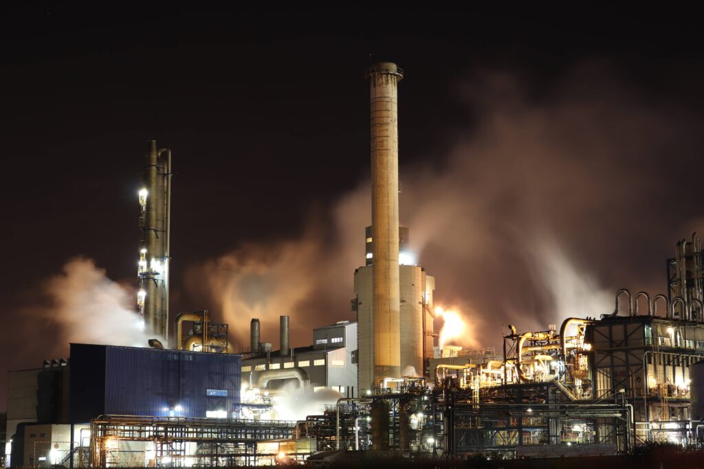 Nighttime industrial refinery with illuminated smokestacks and steam emissions, featuring a complex network of pipes and structures involved in the refining process.