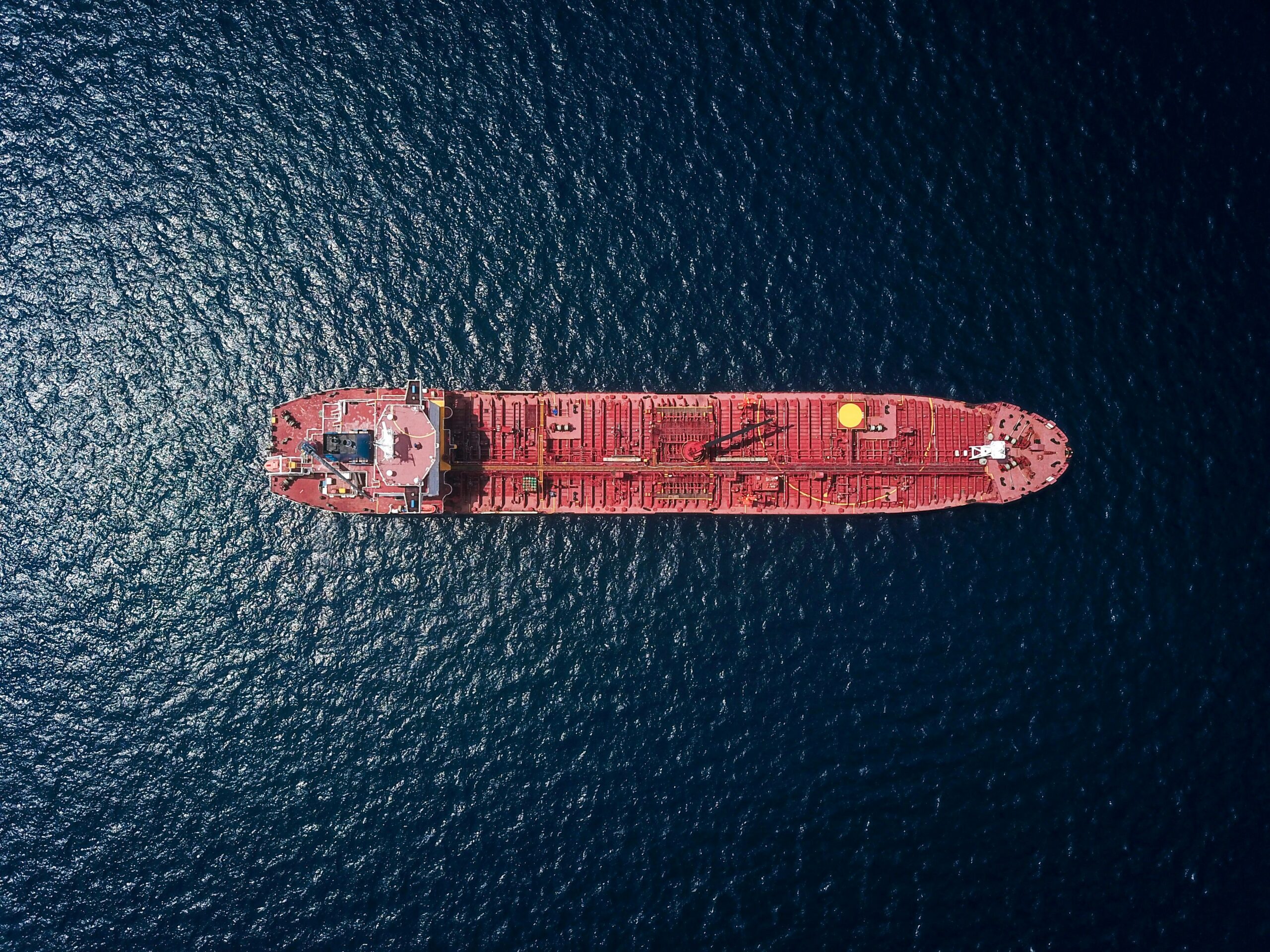 Top-down aerial view of a large red cargo ship navigating through deep blue ocean waters, highlighting the vessel's structure and the surrounding sea.